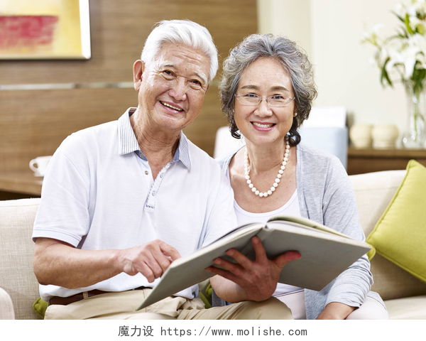 亚洲老年夫妻坐在沙发上拿着书看着镜头微笑高级亚洲夫妇一起阅读一本书